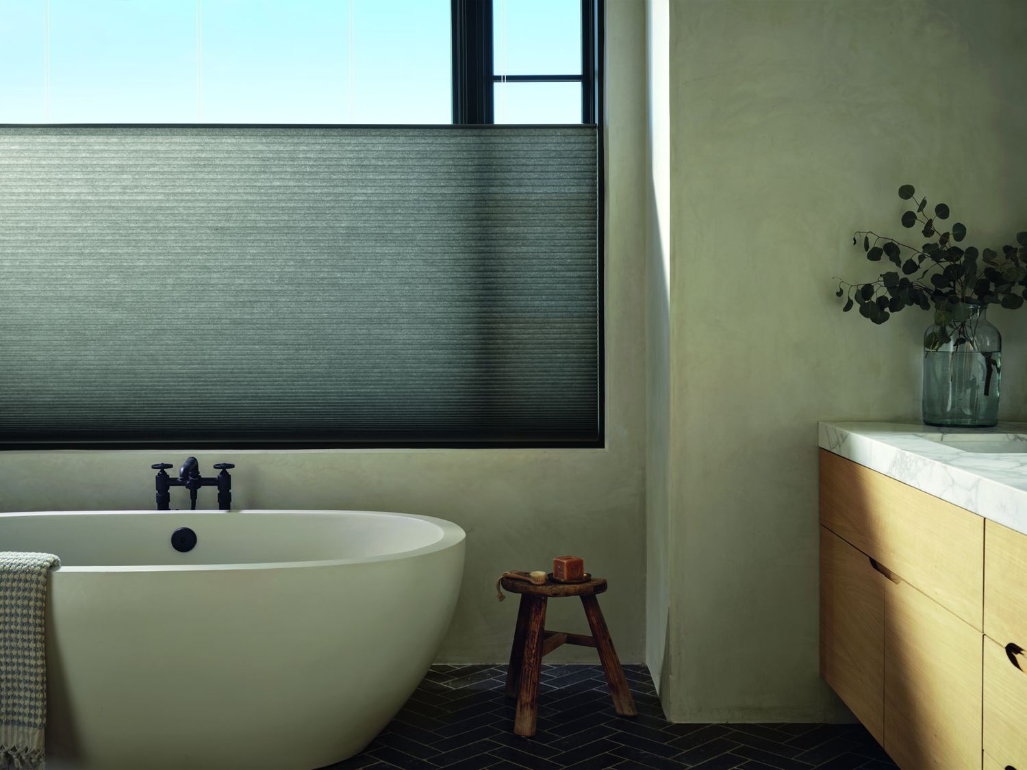 A dark grey honeycomb shade lowered from the top of a bathroom window above a freestanding tub, with daylight coming in above