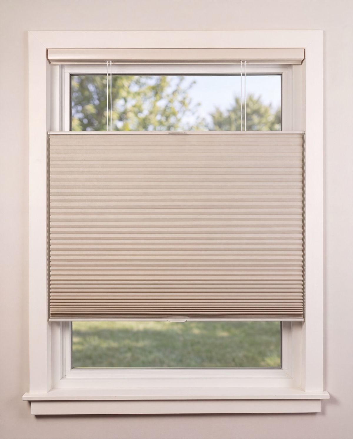 A cream honeycomb shade lowered from the top of a window, with the open glass above it showing green trees and sky